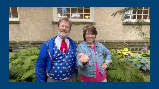 John Hayden, dressed in a outfit of an iconic American figure, and Jeanne Elberfield, smiling and extending a hand, stand outside a Quaker Meeting House, encouraging participation in Philadelphia Yearly Meeting’s (PYM) Granting Groups. PYM is a Quaker non-profit dedicated to philanthropy, stewardship, and supporting the community through grants.