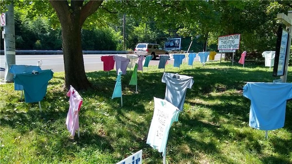 A Memorial to the Lost® installation displays colorful t-shirts on stakes in a grassy area under a tree. Each t-shirt bears the name and death date of an individual lost to gun violence, symbolizing the impact of gun violence in Philadelphia. A sign in the background reads, “Injustice Anywhere Threatens Justice Everywhere.” This powerful memorial serves as a call for peace and gun violence awareness. has context menu