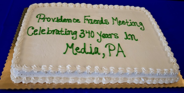 A rectangular white cake with green icing reads “Providence Friends Meeting Celebrating 340 Years in Media, PA.” The cake has decorative white frosting along the edges and is displayed on a gold tray with a blue tablecloth. This photo highlights the 340th-anniversary celebration of Providence Monthly Meeting in Media, Pennsylvania, as described in the event article.