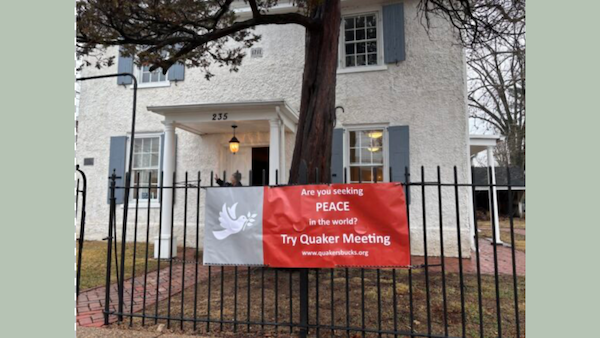 A white historic-style house with blue shutters and a black metal fence in front. A red and white banner is attached to the fence, displaying the message: ‘Are you seeking PEACE in the world? Try Quaker Meeting’ along with a website URL, ‘www.quakerbucks.org.’ The banner features a white dove carrying an olive branch. The house has a porch with two white columns, a black lantern hanging above the entrance, and the number ‘235’ displayed above the door. The setting appears to be on a cloudy day.