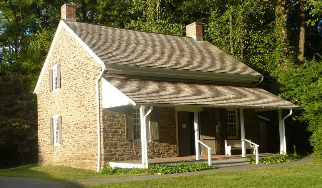 A small, historic stone meetinghouse with a steep, shingled roof and two chimneys, surrounded by trees. The front features a porch with white railings and two windows.